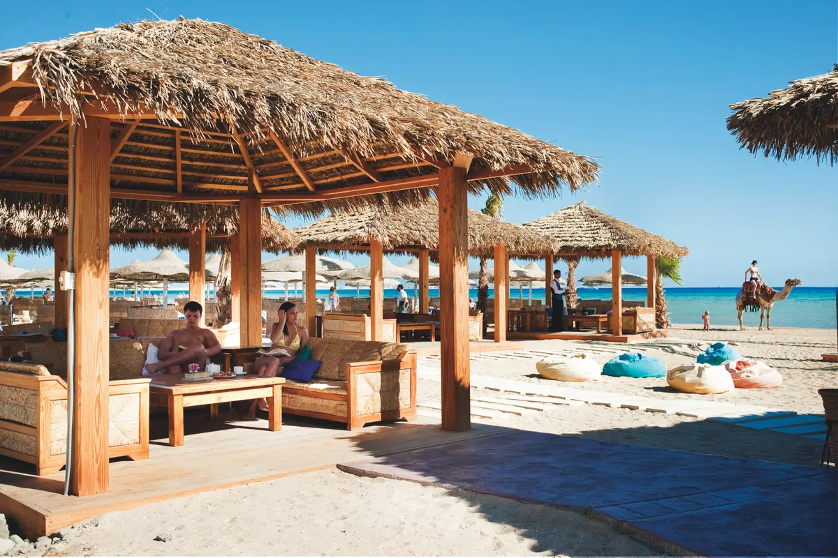 Amwaj Soma Hotel & Spa – Strohgedeckte Strandunterstände mit Holzsitzen, Sitzsäcken und einem Kamel am Ufer