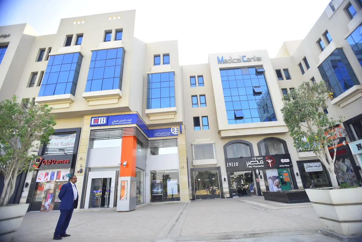 Prima Vista – Medical Office & Commercial Mall – محلات وبنوك في الطابق الأرضي أسفل الواجهة الزجاجية للمركز الطبي مع أشجار الساحة