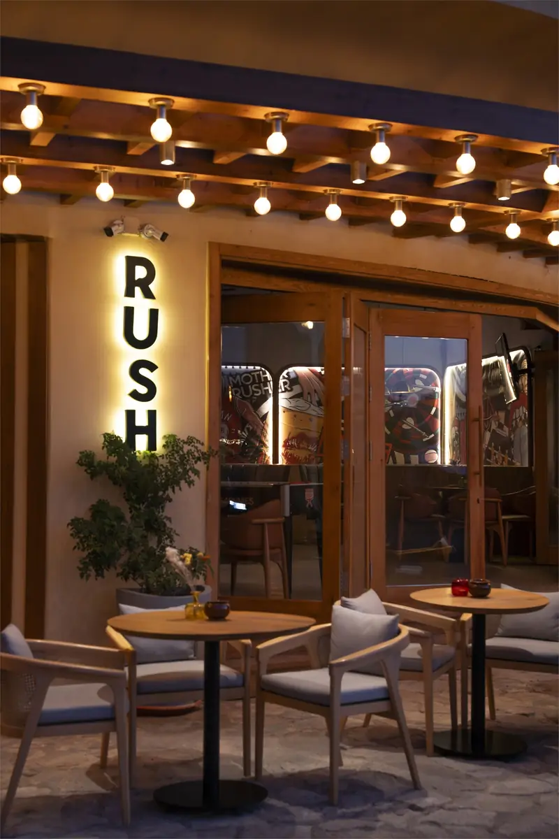 Rush – Sports Bar – مدخل زجاجي بلافتة رأسية مضاءة من الخلف وطاولات صغيرة وسلاسل مصابيح