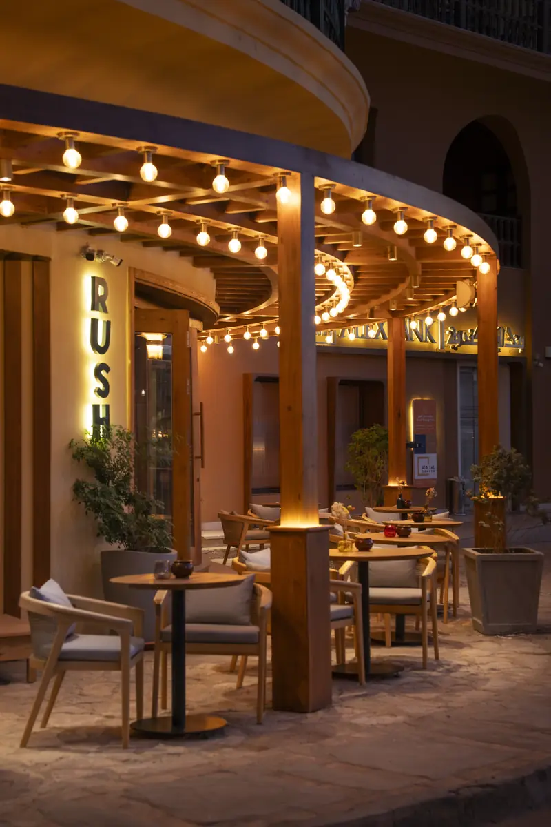 Rush – Sports Bar – أعمدة خشبية مضاءة من الأسفل على امتداد التراس المنحني المضاء بسلاسل المصابيح ليلًا